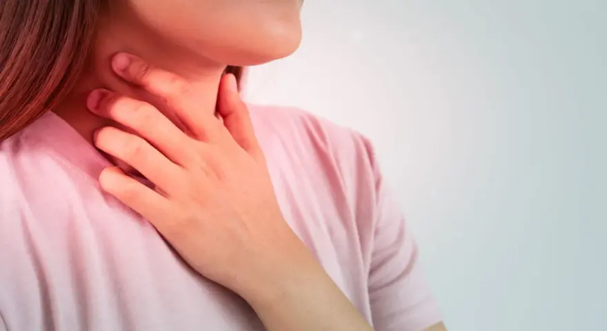 Kobieta z ręką na szyi, odczuwająca ostry ból gardła przy przełykaniu, bez gorączki. Czerwony rumień sugeruje stan zapalny.